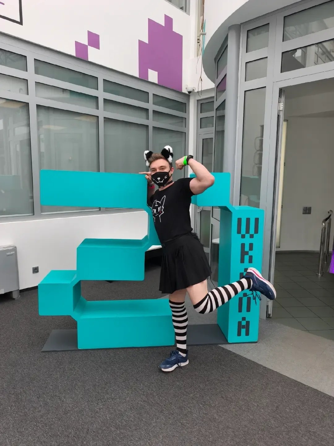 Daniil in a black skirt and striped stockings beside a turquoise “21” installation